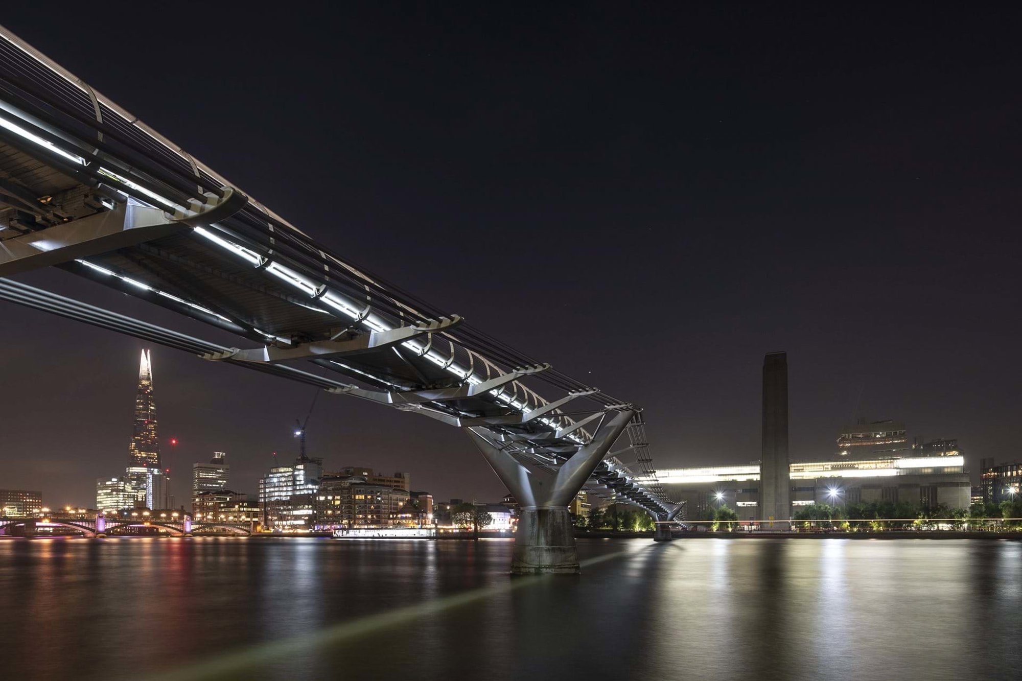 The Millennium Bridge illuminated at night like a ‘blade of light’. The Bridge has a strong axial relationship with St Paul’s Cathedral but avoids replicating the same gesture with the redundant central chimney of the Bankside power station, now the Tate Modern. © Nigel Young / Foster + Partners